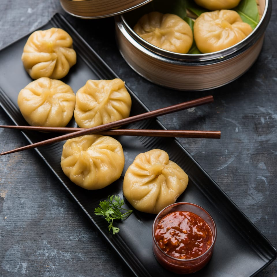Nepalese momos on a black plate with chopsticks, chili sauce, and a bamboo steamer.