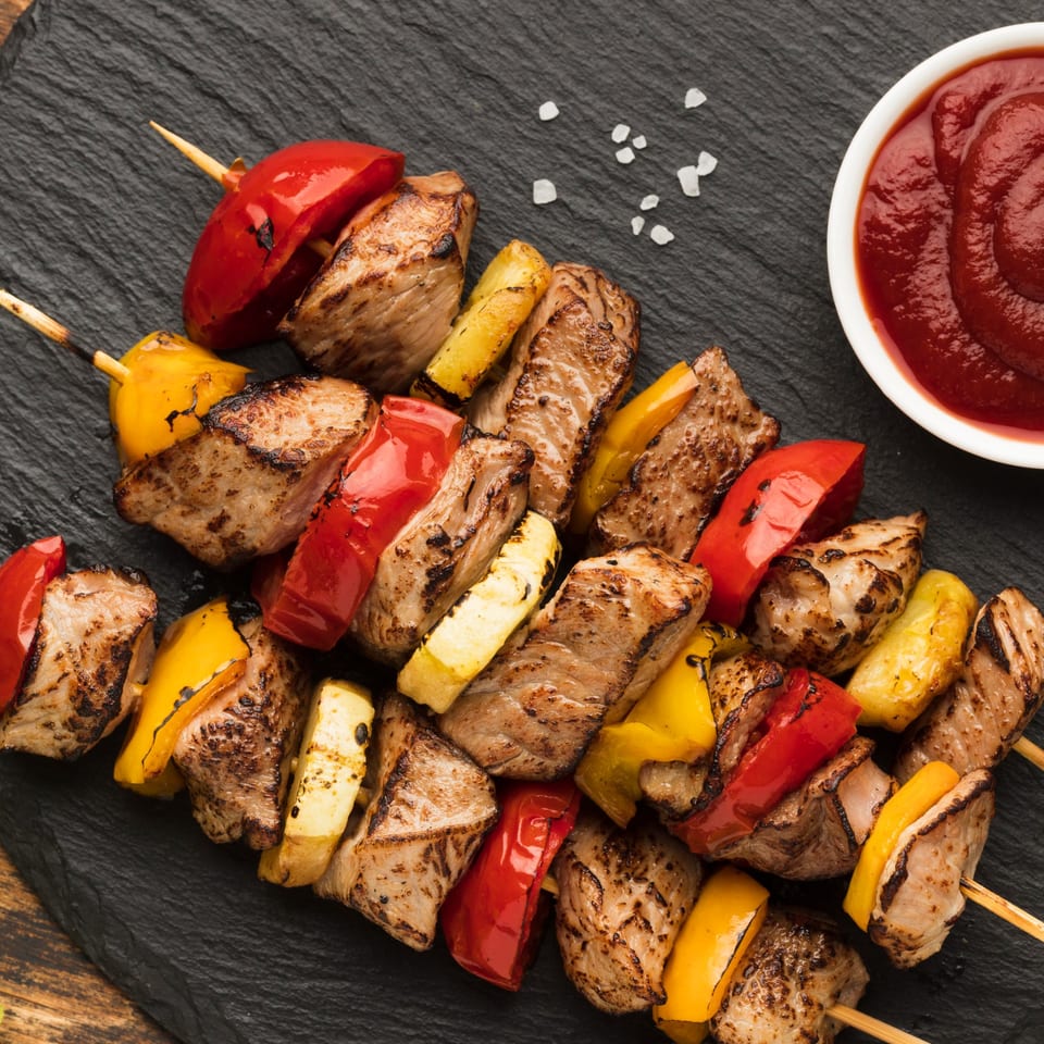 Grilled meat and vegetable skewers with red and yellow bell peppers, served with a side of sauce.