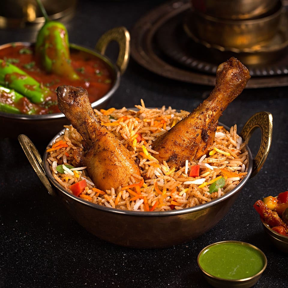 Chicken biryani with two drumsticks in a brass bowl, alongside chili curry and green chutney.